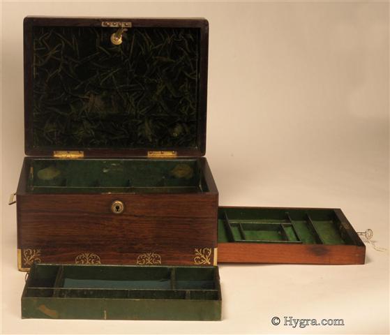 Figured rosewood box with rounded edges in alternating brass and rosewood having fine inlaid brass accents depicting  stylized neo-classical motifs . The box is of high quality, which continues into the compartmentalized interior. There is a lockable side drawer of dovetail construction fitted for jewelry. It is lined in velvet and embossed leather. The box has its original leather covered lift-out tray. There is a document wallet in the lid with a mirror behind.  Both the main lock and the drawer lock are working and have the same key. Circa 1825   Enlarge Picture